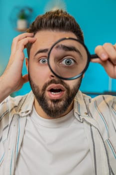 Caucasian young freelancer man holding magnifying glass near face, looking into camera with big zoomed funny big eye, searching, analyzing. Bearded guy sitting at home office table workplace. Vertical