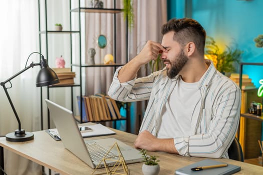 Bored sleepy business man worker working on laptop computer, rubbing eye pinch nose at home office desk. Exhausted tired freelancer workaholic guy eyes pain. Employment occupation workless. Lifestyle