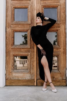 Stylish woman in the city. Fashion photo of a beautiful model in an elegant black dress posing against the backdrop of a building on a city street.