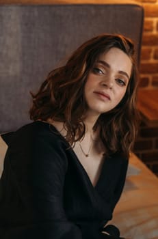 A serene young woman in a black dress sits in a cozy indoor setting, with soft ambient lighting highlighting her features. Vertical frame.