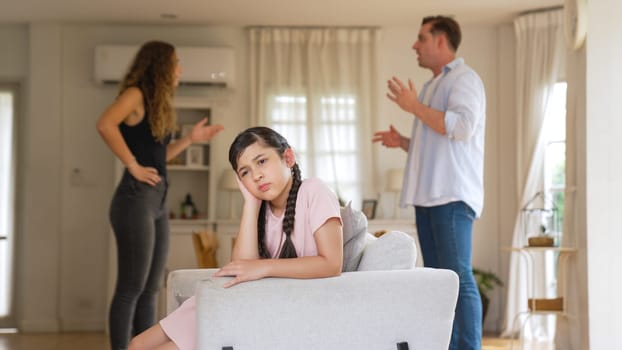 Annoyed and unhappy young girl sitting on sofa trapped in middle of tension by her parent argument in living room. Unhealthy domestic lifestyle and traumatic childhood develop to depression Synchronos