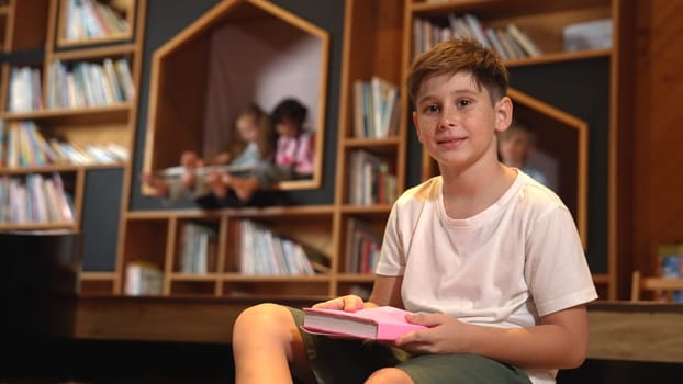 Caucasian boy looking at camera while group of smart students sitting at library. Child studying, learning, reading from novel or textbook while children talking, chitchat about education. Erudition.