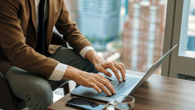 Businessman sitting on furniture working on laptop at ornamented corporate waiting area with cityscape background on the window. Business profession and strategic marketing plan for business success.