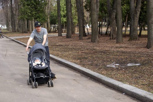 A man is pushing a stroller with a baby in it along a sidewalk. The man is dressed casually, and the baby appears to be looking around curiously. The surroundings are urban, with buildings and pedestrians in the background.