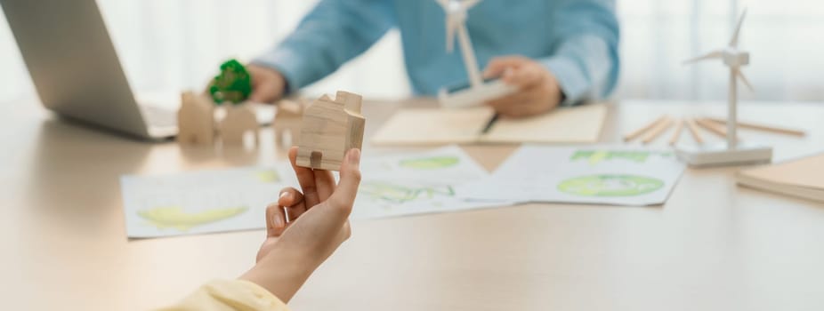 Cropped image of businesswoman presents eco-friendly house by using green design to manager while holding the wooden house block at table with windmill model placed with document. Delineation.