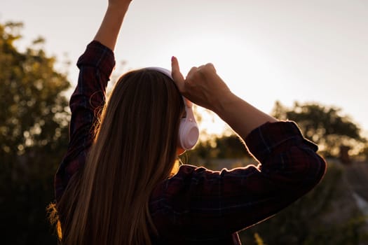 Back view of woman with headphones raising arms against sunset. Outdoor music enjoyment concept with copy space for design and print