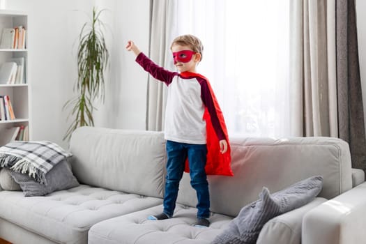 Child in superhero costume with red cape and mask standing on a sofa. Imaginative play and childhood concept. Home interior shot with natural light and space for text