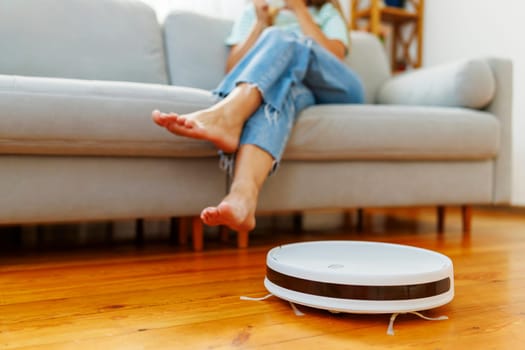 Person relaxing on sofa with feet up while robotic vacuum cleaner works on wooden floor. Home automation and smart cleaning concept