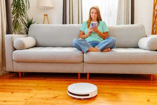 Woman using smartphone on couch with robotic vacuum cleaner on the floor. Modern home and technology concept with copy space. Design for banner, lifestyle blog, and smart home advertisement