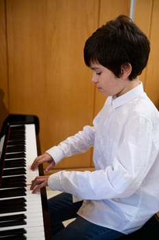 Confident portrait of teen boy in white shirt, musician pianist putting fingers on piano keys while playing piano, enjoying the performance of classical music indoor. Kids education and entertainment.