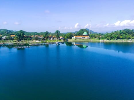 serene lake surrounded by lush green trees, with a white building with a mosque in the background. Around Ranu or Lake Klakah, Lumajang, East Java, Indonesia.