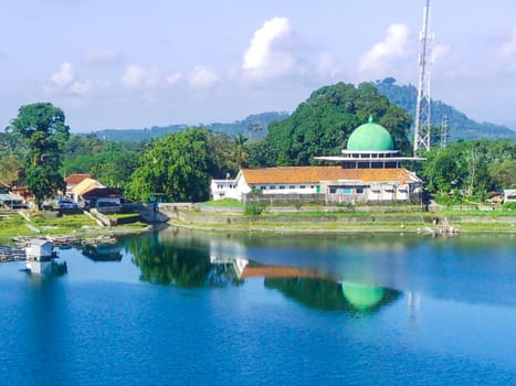 serene lake surrounded by lush green trees, with a white building with a mosque in the background. Around Ranu or Lake Klakah, Lumajang, East Java, Indonesia.