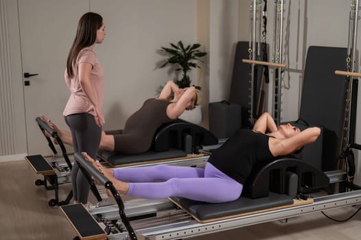 Two pregnant women are doing Pilates on a reformer. The instructor teaches prenatal yoga