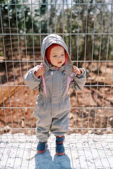 Little girl unzips her hood in overalls while standing near a metal fence. High quality photo