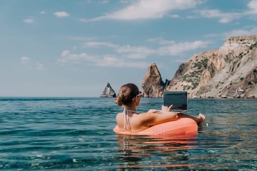 Woman freelancer works on laptop swimming in sea on pink inflatable ring. Pretty lady typing on computer while floating in the sea on inflatable donut at sunset. Freelance, remote work on vacation