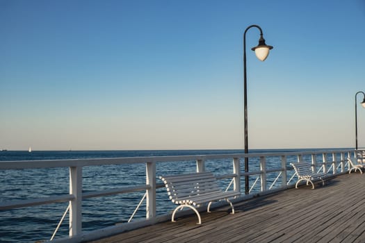 White Old wood bridge pier against beautiful sunset sky natural background, backdrop wallpaper multipurpose sea scene. White benches no people in Gdynia Orlowo, Poland. Wooden pier, molo with marina and beach Touristic destination