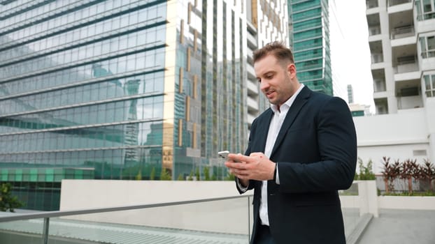 Happy businessman using phone to chatting with team while standing at urban city. Front view of project manager working on telephone with skyscraper view while communicate with marketing team. Urbane.