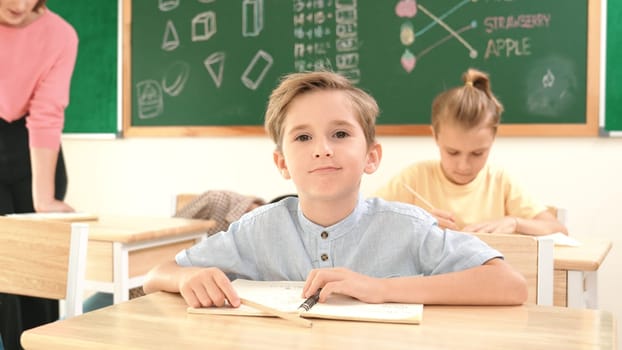 Attractive caucasian boy looking at camera while studying math at school. Diverse cute student doing test while teacher asking asian child question and point at paper in front of blackboard. Pedagogy.