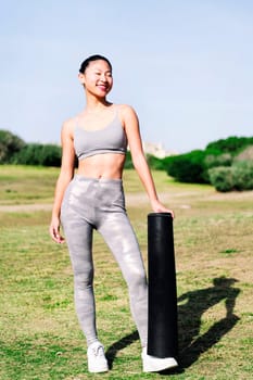 smiling young asian woman in sportswear ready with her mat for a yoga session on the grass in the park, healthy and active lifestyle concept