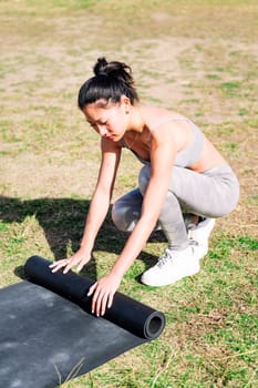 young asian woman in sportswear rolling her mat for a yoga session on the grass in the park, healthy and active lifestyle concept