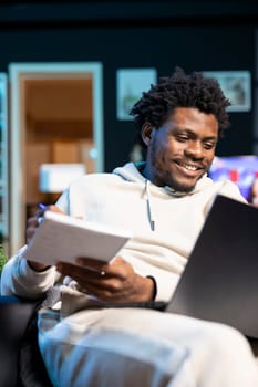 Entrepreneur working from neon lit apartment, writing down company ideas while watching video on laptop. BIPOC man feeling delighted after coming up with startup proposal, using pen to note plan