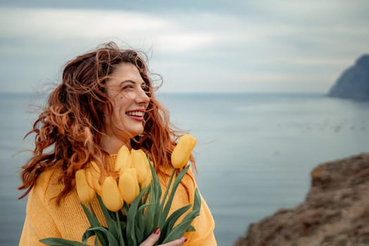 Portrait of a happy woman with hair flying in the wind against the backdrop of mountains and sea. Holding a bouquet of yellow tulips in her hands, wearing a yellow sweater.