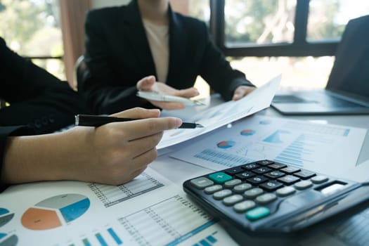 Two people are sitting at a table with a calculator and a stack of papers. One of the people is holding a pen and looking at the papers. The atmosphere seems to be focused and serious