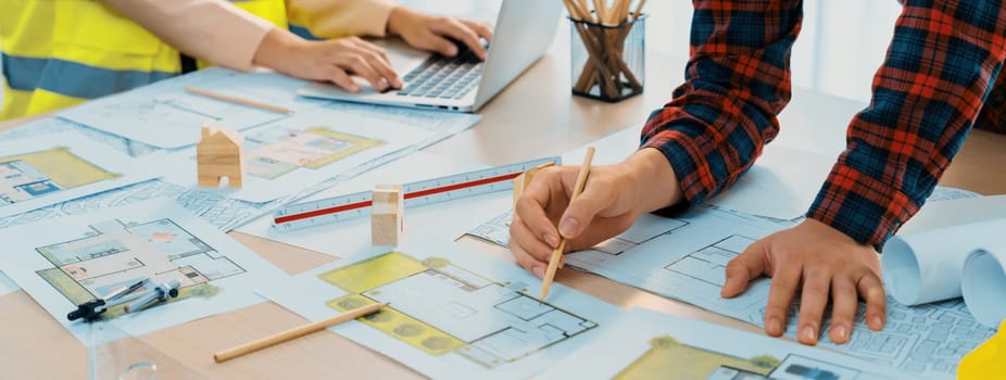 A cropped portrait of professional architect working with blueprint during safety engineer using laptop analysis data at meeting table with document scatter around. Close up. Delineation.