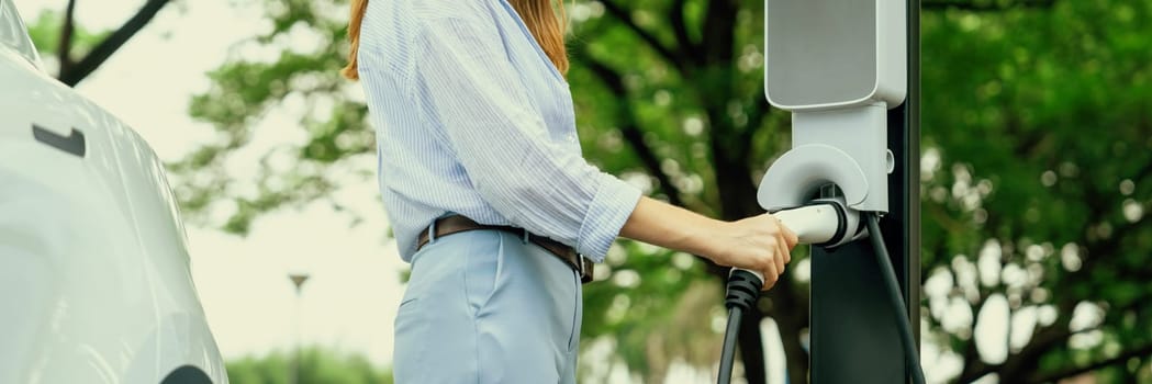 Young woman recharging battery for electric car during road trip travel EV car in natural forest or national park. Eco friendly travel during vacation and holiday. Panorama banner Exalt