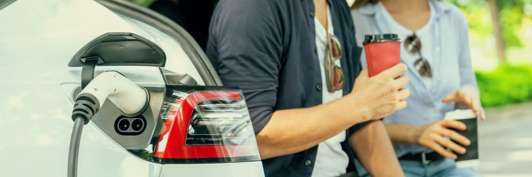 Panorama banner of couple drinking coffee while recharge battery for electric car during road trip travel EV car in greenery forest or national park. Eco friendly travel on vacation and holiday. Exalt