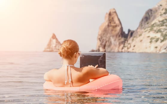 Woman freelancer works on laptop swimming in sea on pink inflatable ring. Pretty lady typing on computer while floating in the sea on inflatable donut at sunset. Freelance, remote work on vacation