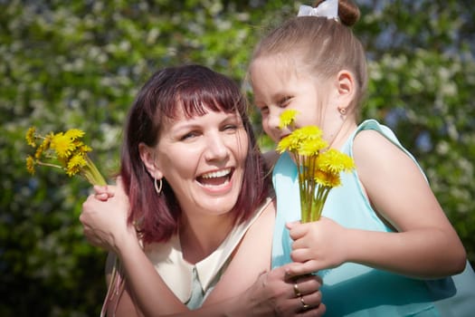 Happy mother and daughter enjoying rest, playing and fun on nature on a green lawn with dandelions and blooming apple tree on background. Woman and girl resting outdoors in summer and spring day