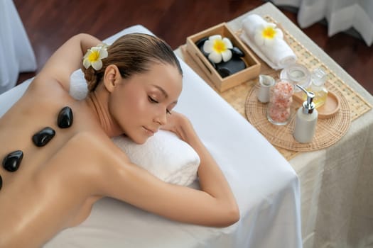 Hot stone massage at spa salon in luxury resort with day light serenity ambient, blissful woman customer enjoying spa basalt stone massage glide over body with soothing warmth. Quiescent