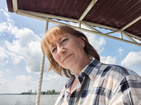 Happy cheerful middle aged woman taking selfie on nature outdoors and sun with water of lake or river on the background in summer day