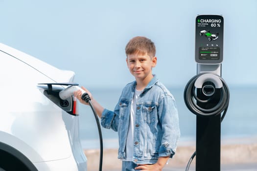 Little boy recharging eco-friendly electric car from EV charging station. EV car road trip travel by the seashore by alternative vehicle powered by clean renewable and sustainable energy. Perpetual