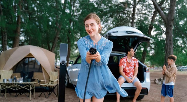 Outdoor adventure and family vacation camping at sea travel by eco friendly car. Cheerful woman or mother holding, pointing EV charger point with playful and happiness posture in campsite. Perpetual