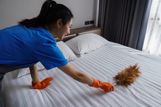 A woman is cleaning a bed with a feather duster. She is wearing a blue shirt and orange gloves