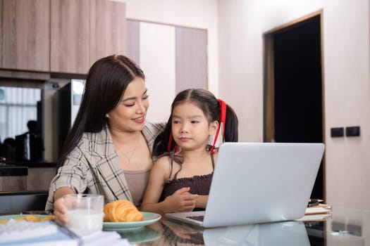 a single mother, sits at home working on a laptop with her daughter beside her watching and encouraging her..