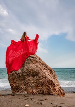 woman sea red dress. Woman with long hair on a sunny seashore in a red flowing dress, back view, silk fabric waving in the wind. Against the backdrop of the blue sky and mountains on the seashore