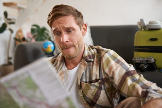 Portrait of man looking confused at travel map, choosing route, going on holiday, preparing for vacation, sitting in living room with suitcase.