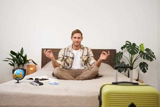 Portrait of young tourist, relaxed smiling man meditating on his bed, sitting in yoga zen pose, has globe and camera, packed suitcase and feels relieved, going on holiday. Mindfulness and tourism.