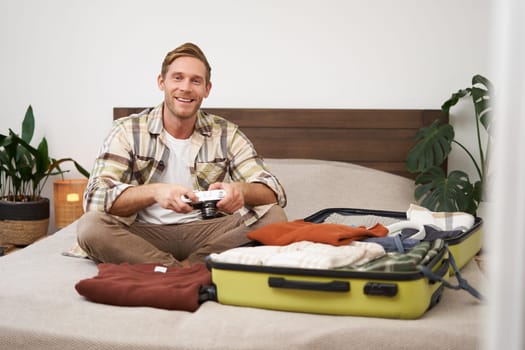 Portrait of young excited man, sits on bed with suitcase, going on holiday, holding digital camera, going through photo album on gadget, preparing for summer vacation.