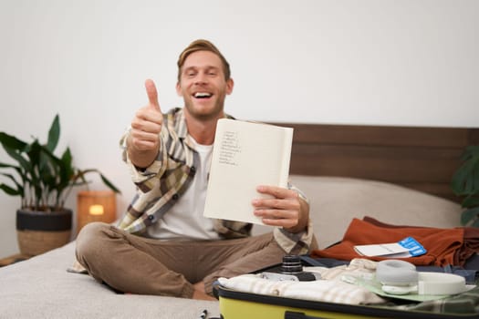 Cheerful young man, tourist with check list, showing his notes in notebook, thumbs up, packing suitcase, going on holiday, travelling abroad and preparing luggage. Copy space