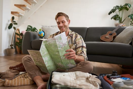 Portrait of handsome young man, traveller with suitcase, looking at paper map, choosing where to go on vacation, going on holiday, choosing route.