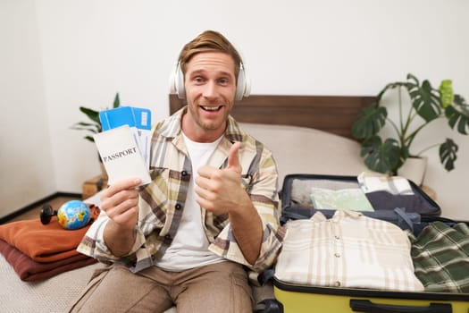 Portrait of smiling man, tourist shows two tickets and passport, goes on holiday, traveller makes thumb up in approval, like travel agency. Copy space