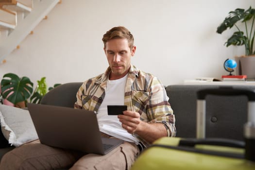 Portrait of cheerful man, going on holiday, plans a travel journey abroad, has laptop opened, sits in living room with suitcase, books a hotel.