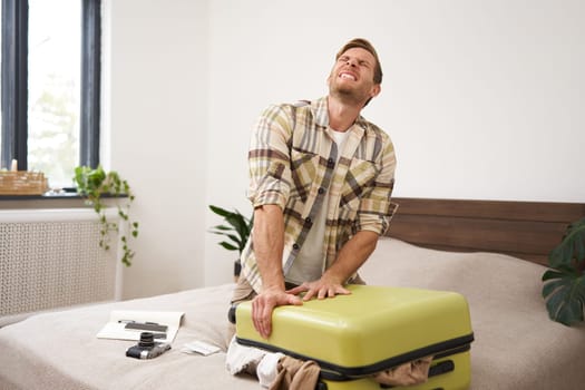 Portrait of young man sweating, shutting his suitcase, trying to close the luggage with lots of clothes, sitting on bed, packing things for summer vacation, going on holiday or business trip.