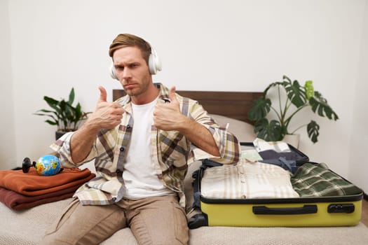 Portrait of handsome man in wireless headphones, shows thumbs up, sits with suitcase, listens to music and packs clothes for vacation, goes on holiday. Copy space