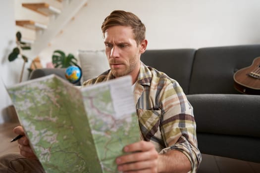 Portrait of happy young man, tourist with suitcase, sits in living room and looks at map, chooses holiday destination, picks route for travelling.