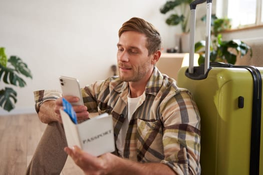 Close up portrait of handsome young man, sits on suitcase, holds passport, flight tickets and his mobile phone, using smartphone app to check departure time.